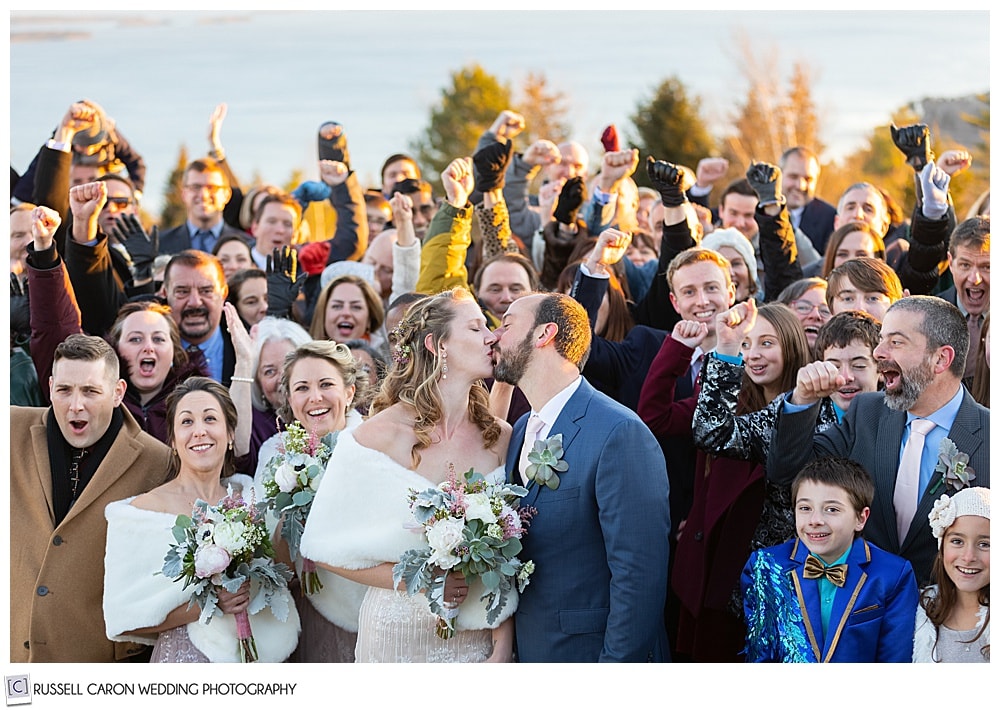 bride and groom share a joyful wedding kiss surrounded by their families and friends