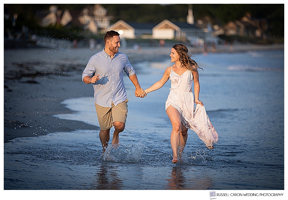 Beachy Kennebunk engagement image of man and woman holding hands and running in the ocean