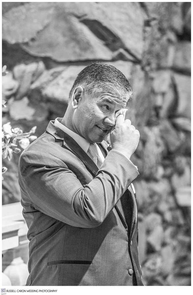 black and white photo of a groom wiping his eyes as his bride walks toward him