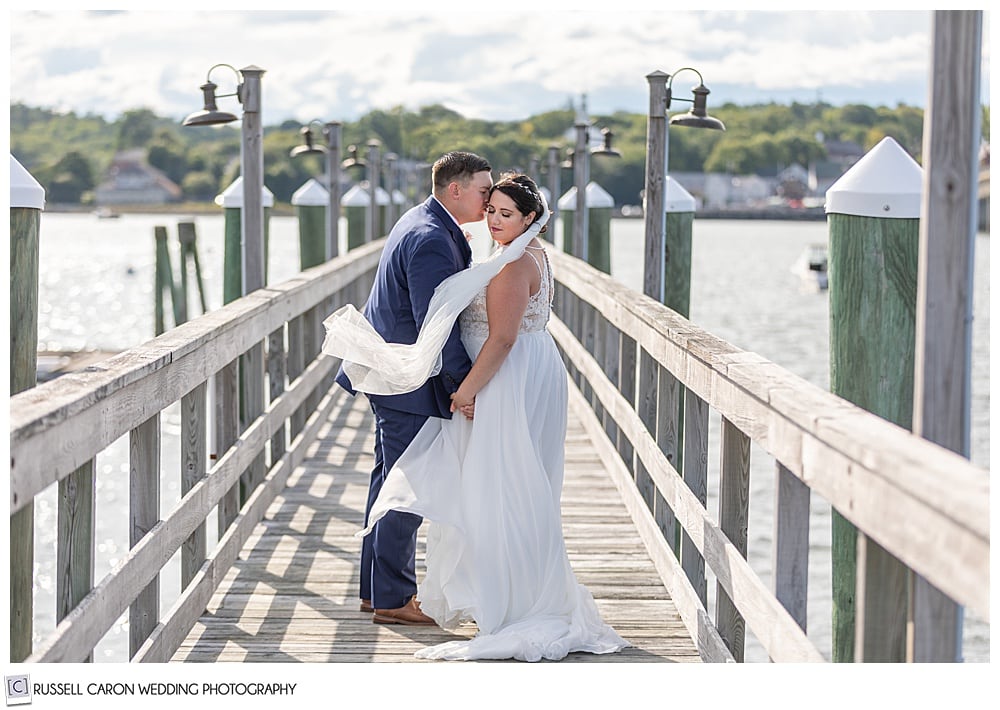 bride and groom on a dock with the groom kissing the bride's temple