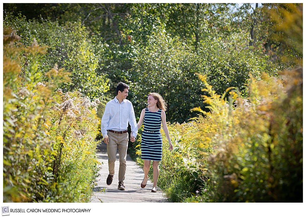 man and woman walking hand in hand on a boardwalk, they're looking at each other