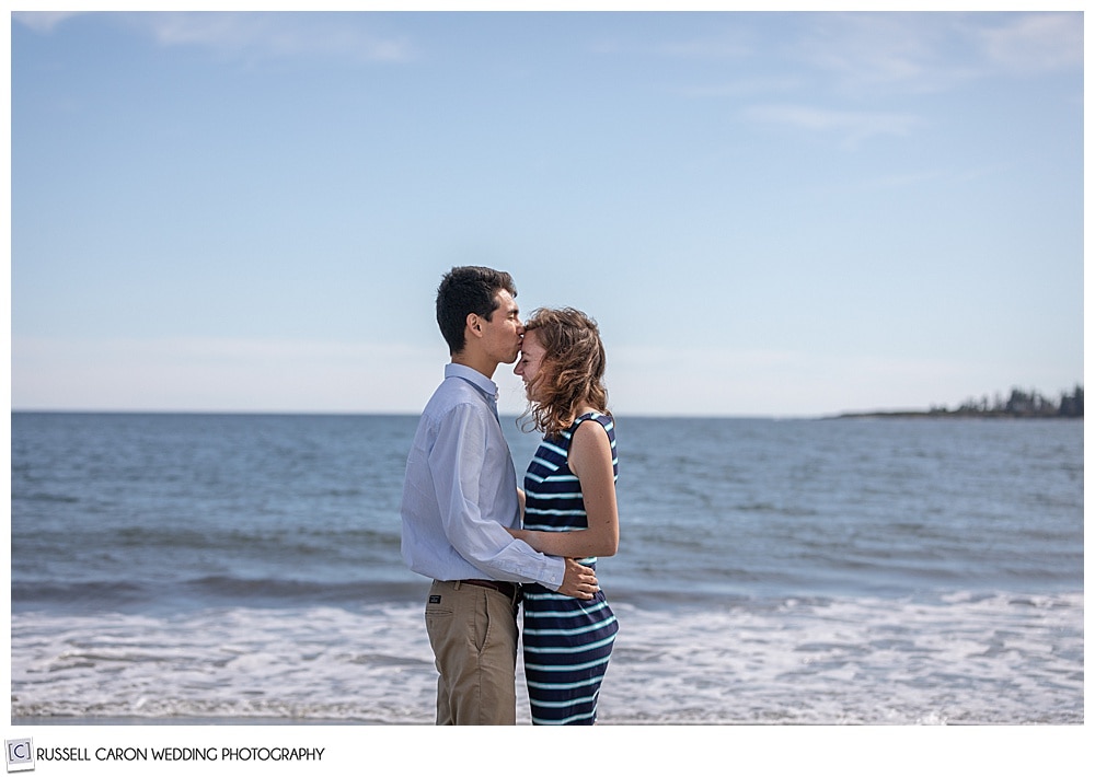 man and woman, standing face to face by the ocean, the man is kissing the woman on the forehead, during their fun crescent beach engagement session, Cape Elizabeth, Maine