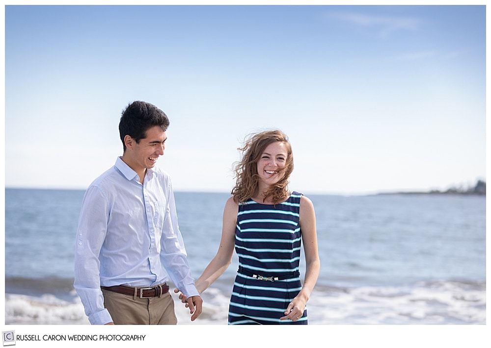 man and woman, holding hands, laughing by the ocean, the woman is looking at the camera