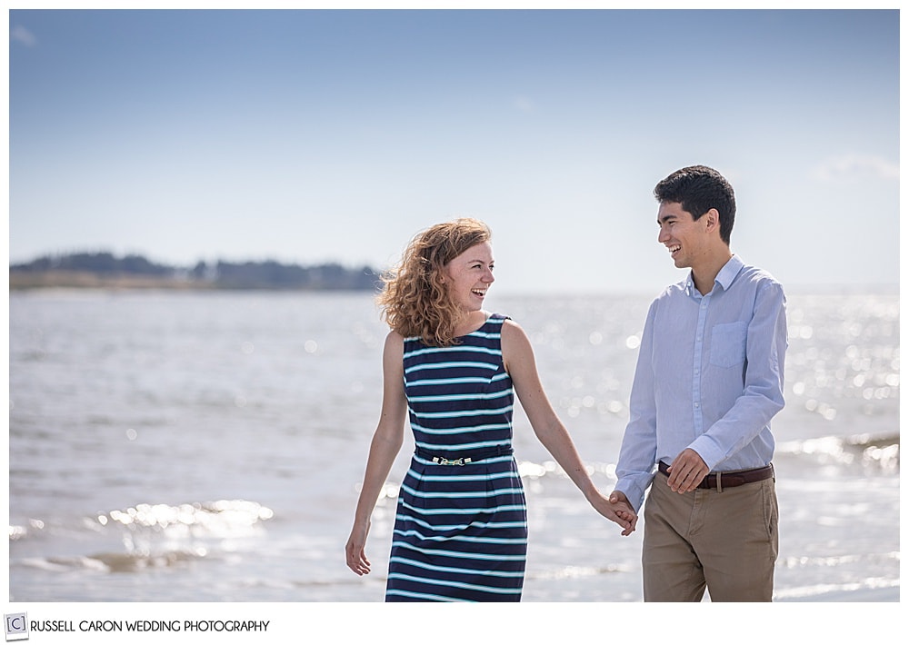 man and woman holding hands, smiling at each other, as they walk along on their fun crescent beach engagement session, cape elizabeth, maine