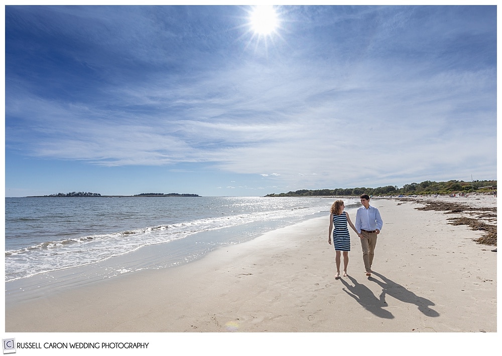 man and woman holding hands and walking on crescent beach, Cape Elizabeth, during their fun crescent beach engagement session