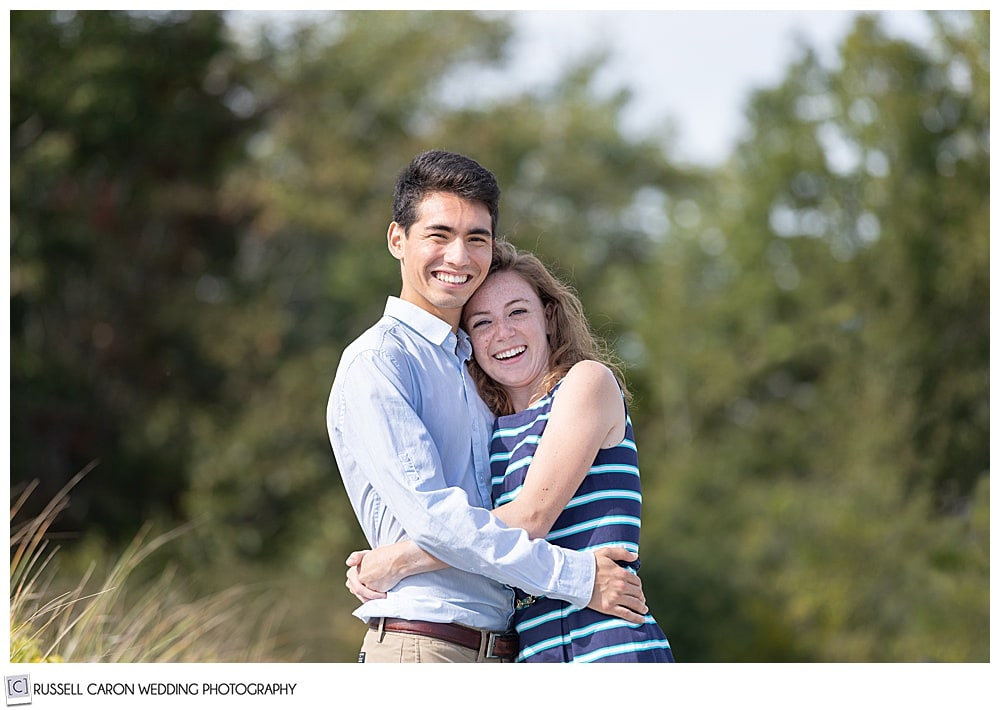 man and woman, near the woods, hugging each other, smiling at the camera during their fun crescent beach engagement session, cape elizabeth, maine