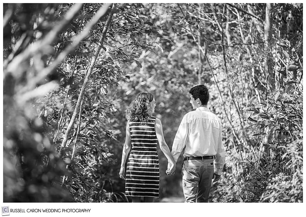 black and white photo of a man and woman, holding hands, walking in the woods