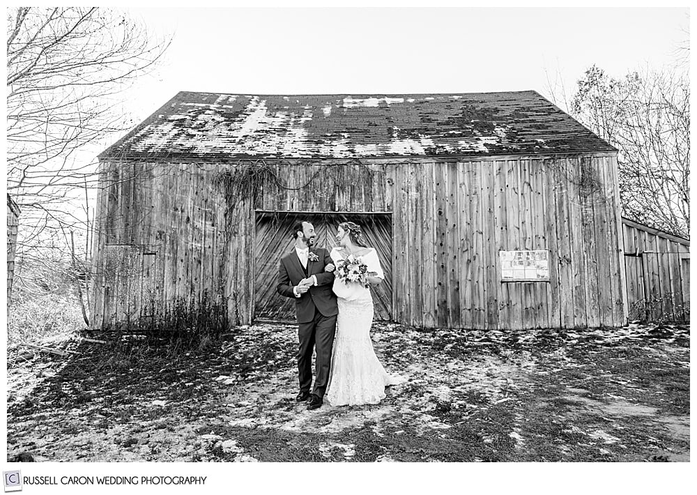 black and white photo of a bride and groom walking side by side in front of an old barn