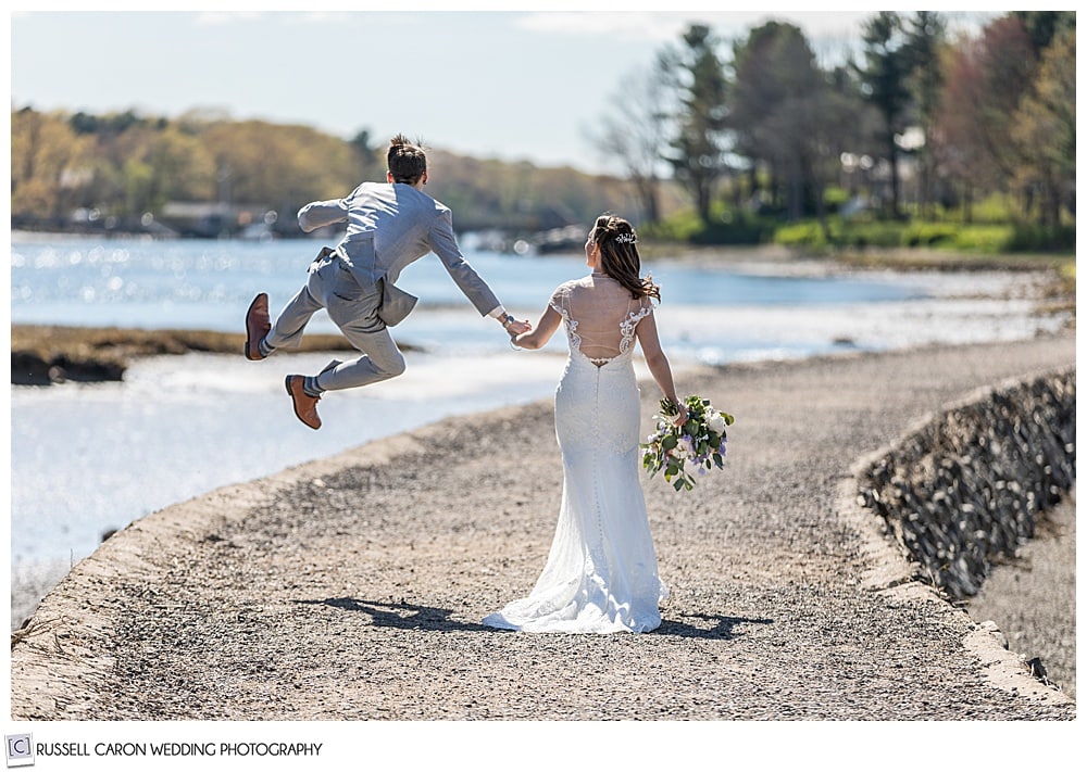 bride and groom walking hand-in-hand, away from the camera, on a path near the water, while the groom jumps and clicks his heels together