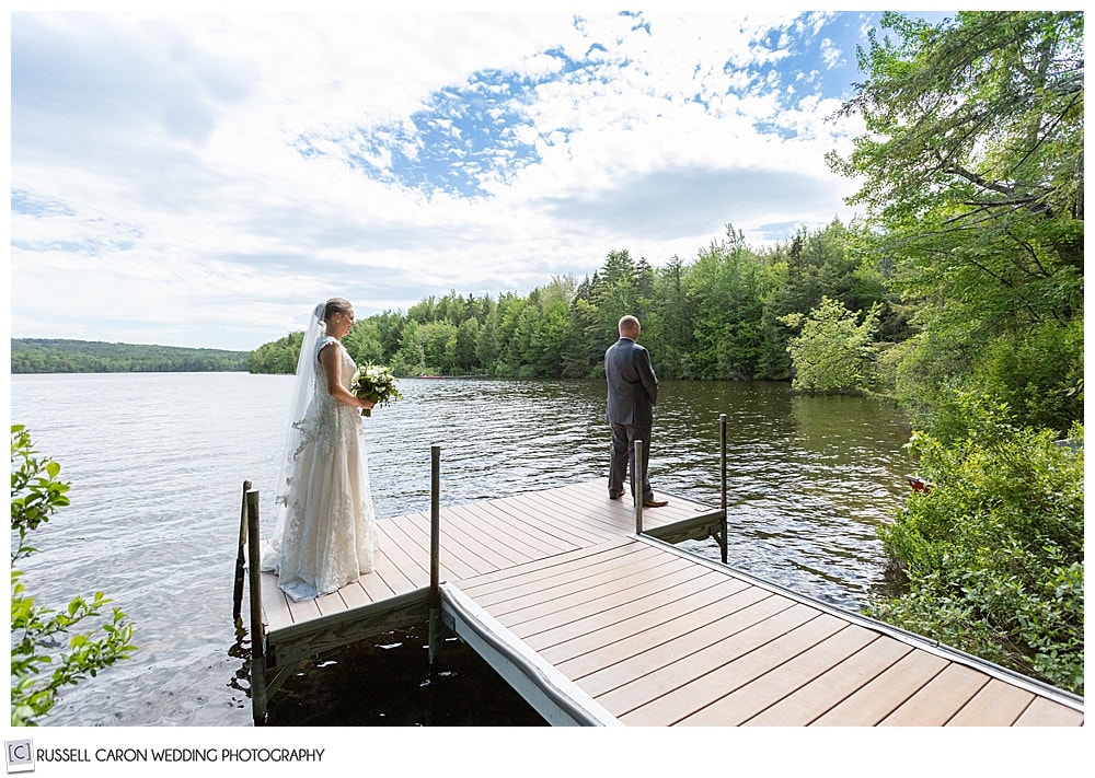 groom stands on a dock with his back to his bride