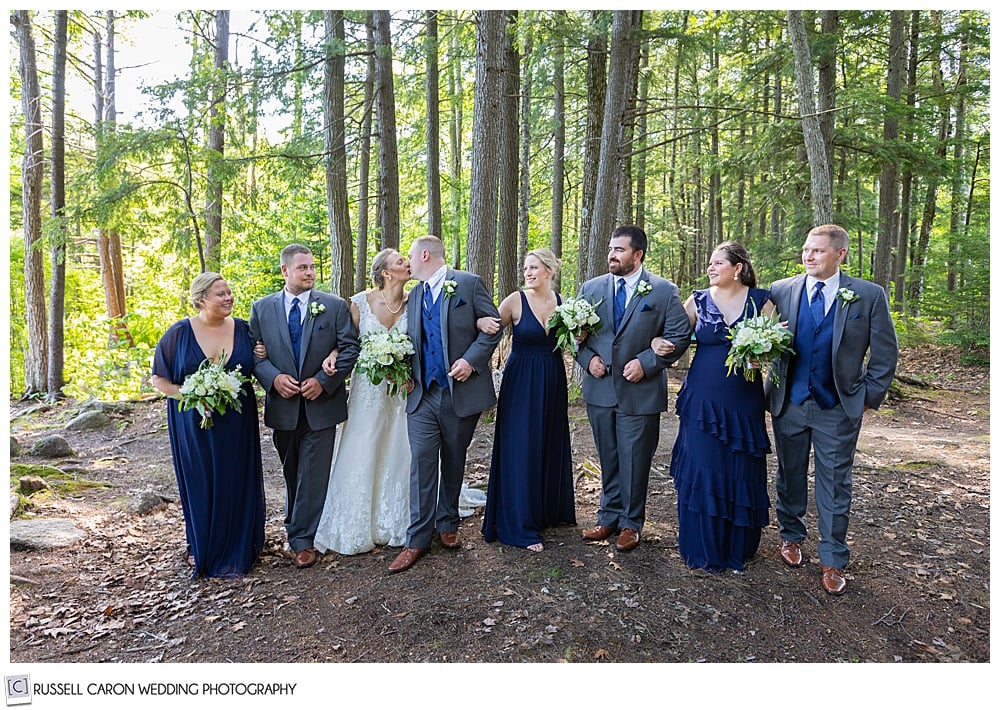 bridal party walking in the woods, arm in arm, while bride and groom kiss