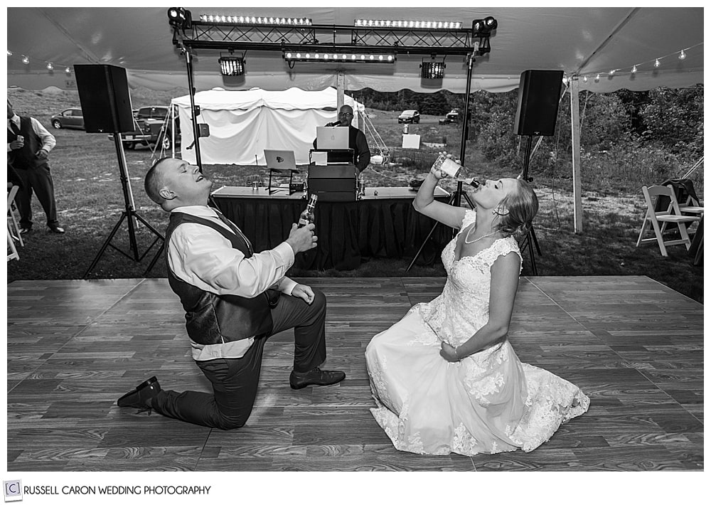 black and white photo of bride and groom, each with one knee on the dance floor, chugging a beverage