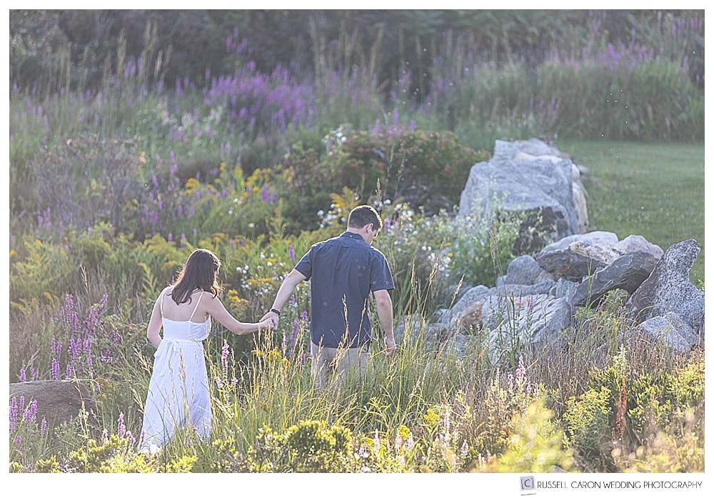 man and woman walking in a field of flowers at their Nubble Light engagement session