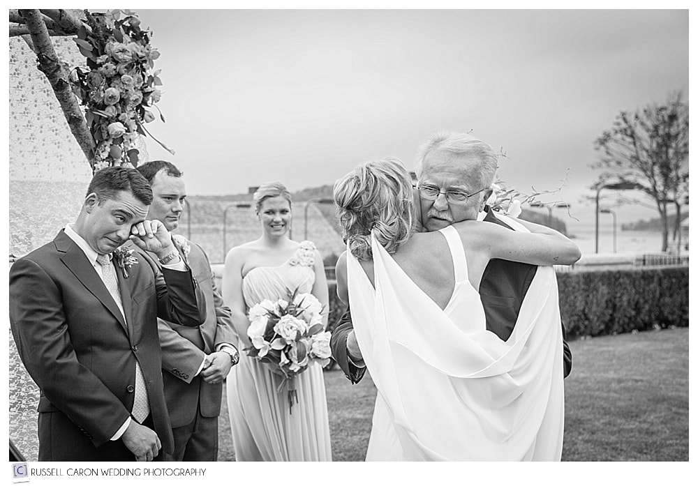 Bride hugging her father, as her groom wipes a tear
