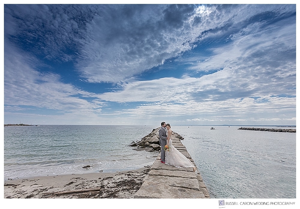 bride and groom standing on the Kennebunkport Maine Breakwater, with dramatic blue skies with white clouds