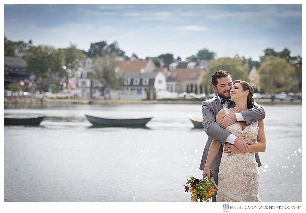 groom hugging bride from behind, on the banks of the Kennebunk River, with three dories 