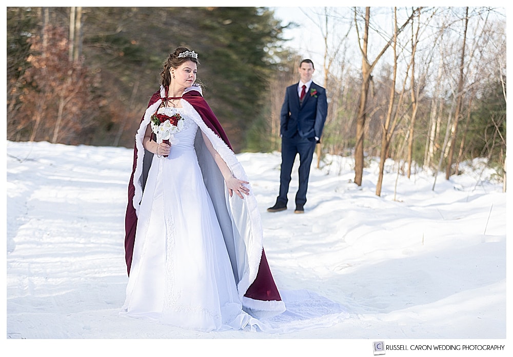 groom looking at bride who's wearing a red cape during a wintery Maine wedding