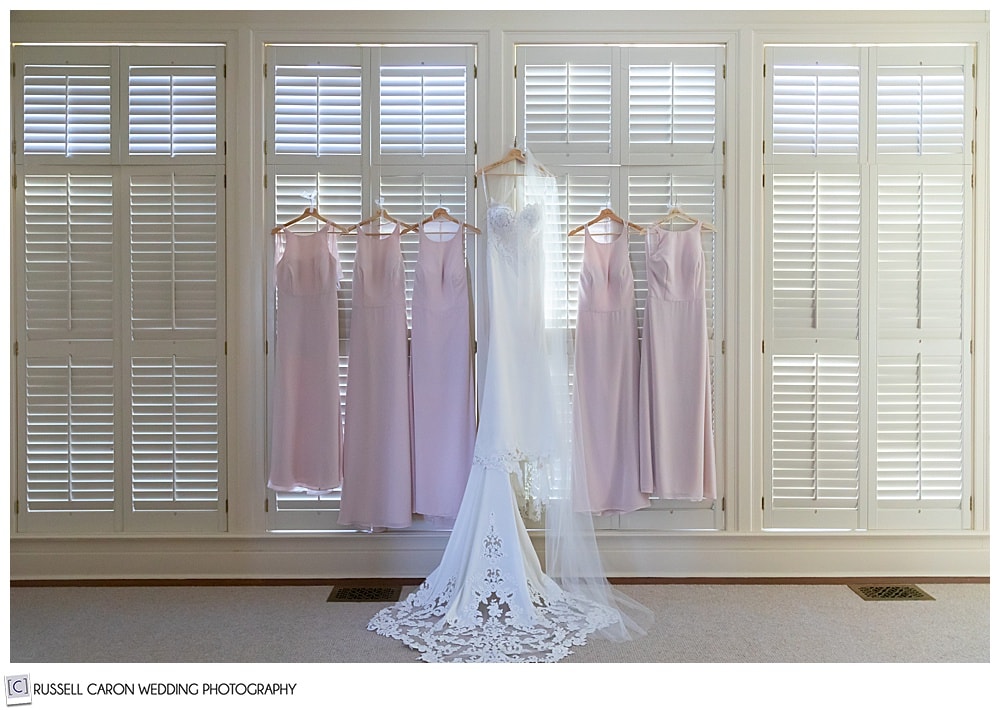 bride's white wedding dress hanging on levered shutters with the pink bridesmaid's dresses