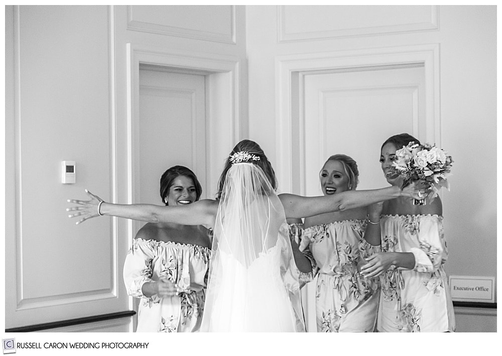 black and white photo of a bride, with her back to the camera, arms outstretched to hug her bridesmaids, who are smiling at her