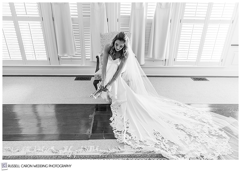 black and white photo of bride, with long white dress and veil, sitting in a chair, putting on her shoe