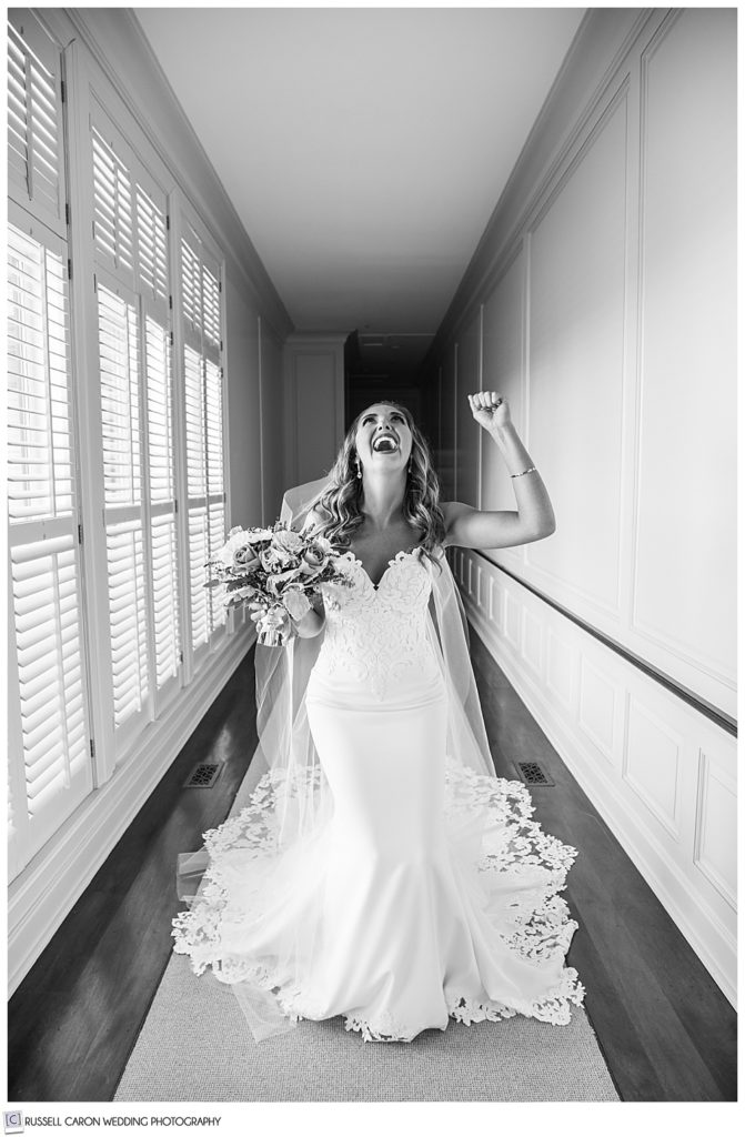 black and white photo of a bride dressed in a long white dress and veil, throwing her head back, and pumping her fist, in a hallway