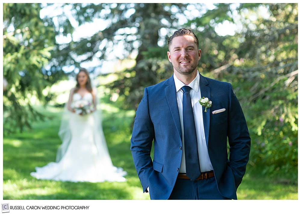 groom dressed in blue suit and tie, with his back to his bride, waiting to see her for the first time