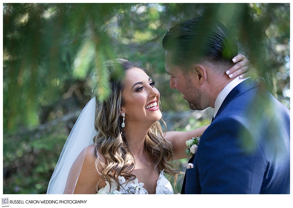 bride, her hand around the groom's neck, smiles during their first look in a grove of trees