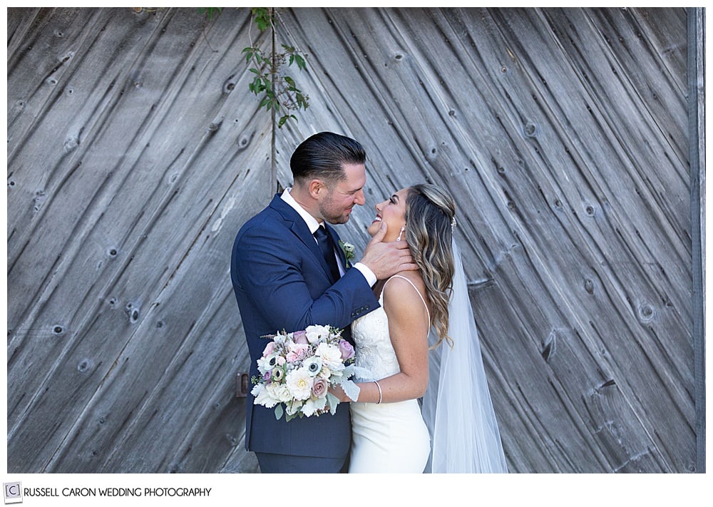 bride and groom standing in front of a barn, groom has his hand on the bride's face as he goes in for a kiss