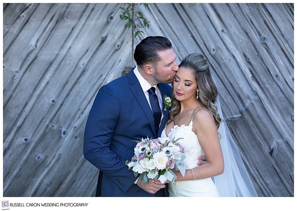 bride and groom standing in front of an old barn, bride's eyes are closed, as groom kisses her on the temple