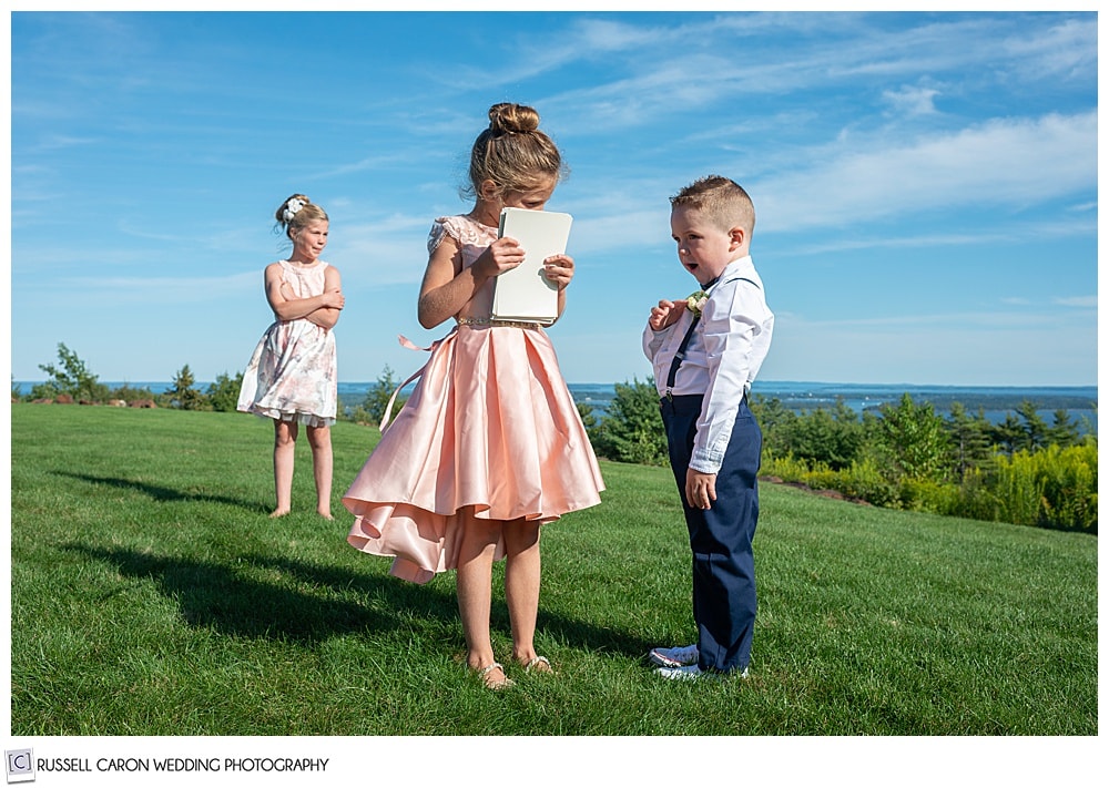 two girls in dresses, and a boy in a white shirt, blue pants and suspenders, play on a grassy hill