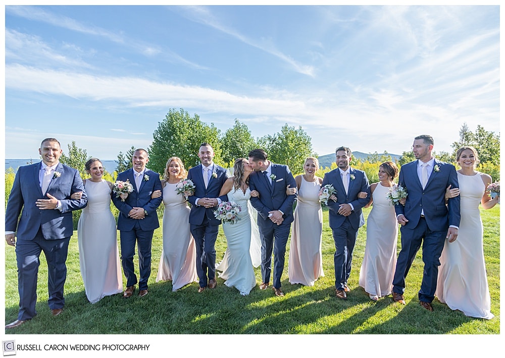 bridal party walking arm in arm, bride and groom kissing, while bridal party looks on