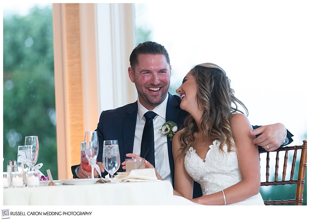 bride and groom sitting next to each other, groom has his arm around bride, she has turned toward him, they're both smiling