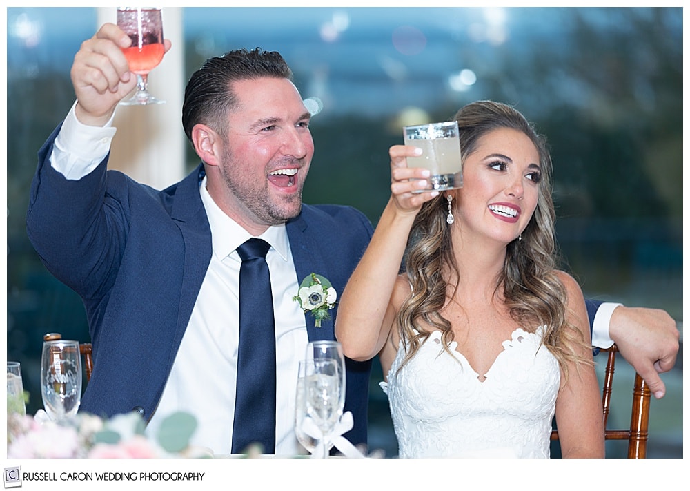 bride and groom, sitting next to one another, raising their glasses in a toast, and smiling