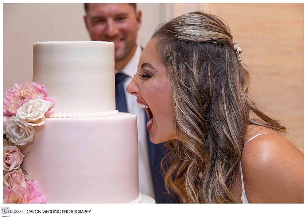 bride pretending to take a bite out of the entire wedding cake, groom is in the background smiling