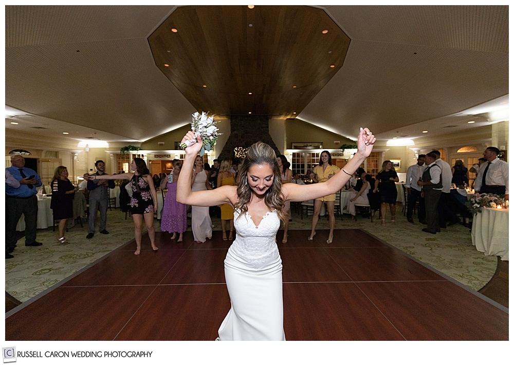 bride in the foreground, holding a bouquet in one hand, while single ladies gather behind