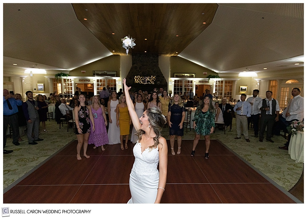 bride in the foreground, facing the camera, but turning as she lets go of her bouquet, to the ladies waiting behind her