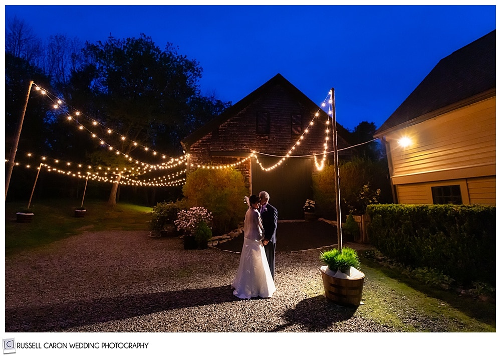 bride and groom standing in front of a barn with twinkly lights in the dark