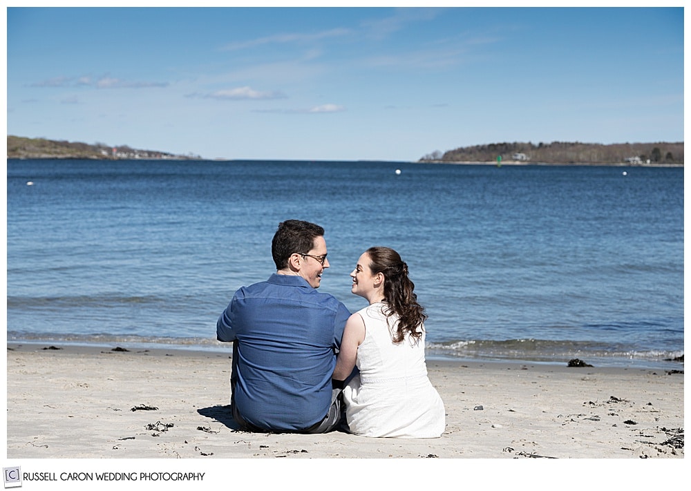 a couple is sitting in the sand at a willard beach engagement session, south portland, maine