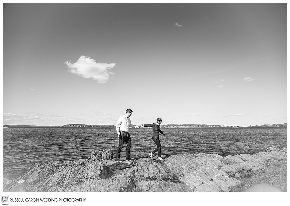 a black and white photo of a man and woman waling on the rocks at the coast of maine