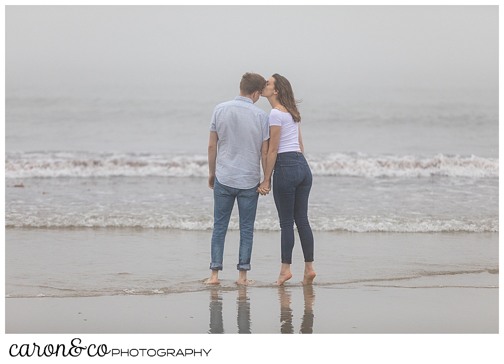 dreamy Maine engagement photo of a man and woman, their backs to the camera, as they kiss at the edge of the ocean