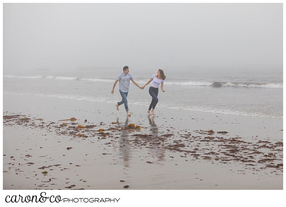 man and woman running in the fog at Biddeford Pool Beach, during a misty, dreamy maine engagement photo session