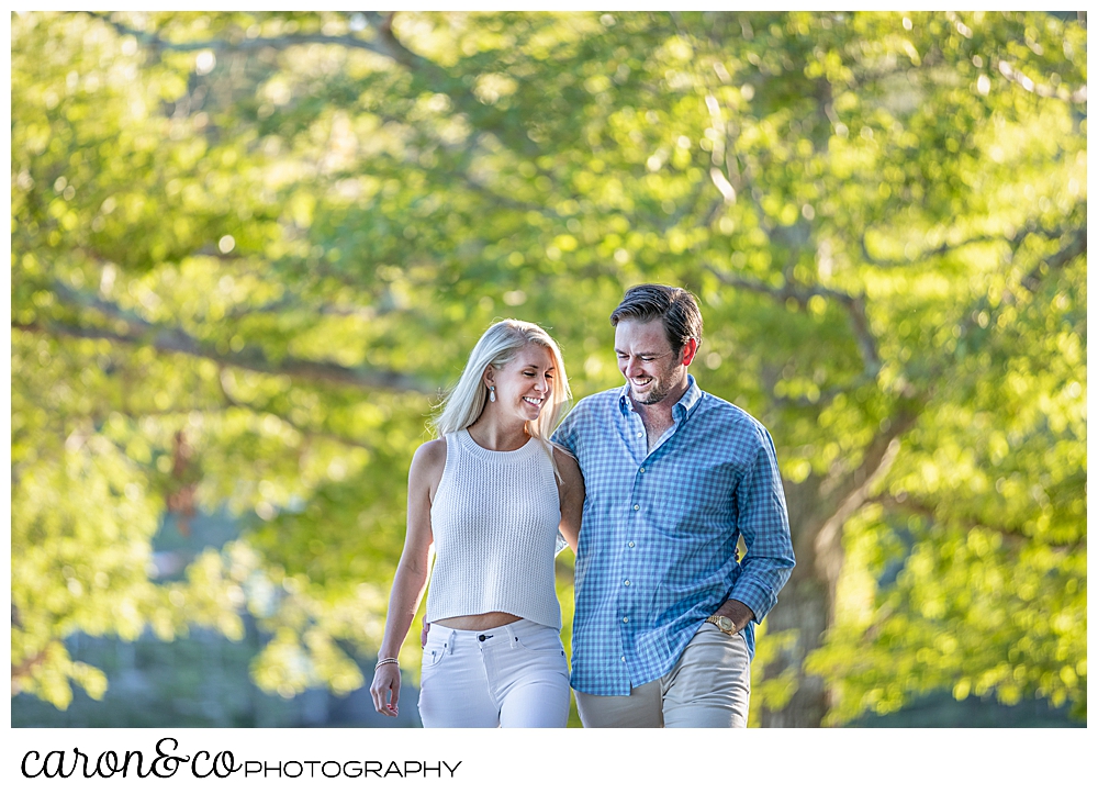 A blonde woman wearing a white top and pants, and a man wearing a blue shirt and tan pants, are walking in a park with their arms around each other