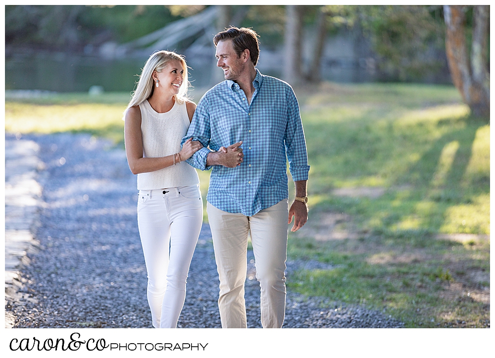 a woman wearing white, and a man wearing a blue shirt and tan pants, are walking arm in arm on the shore during a Boothbay Harbor engagement photo session