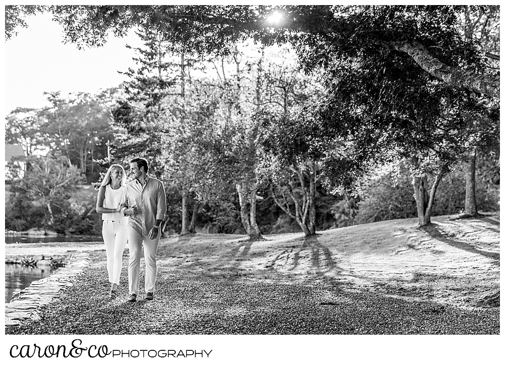 a black and white photo of a man and woman walking in a park along the water's edge, during a Boothbay harbor Maine engagement session