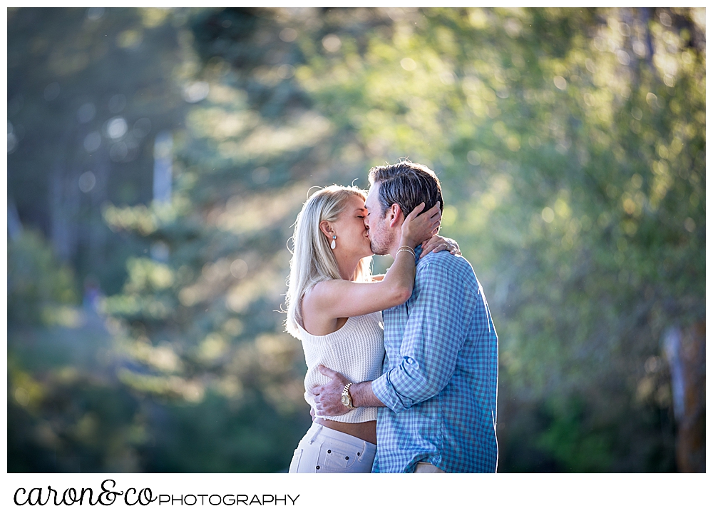 A woman in white and a man in blue and tan, are kissing in a park during a Boothbay Harbor engagement session in Maine