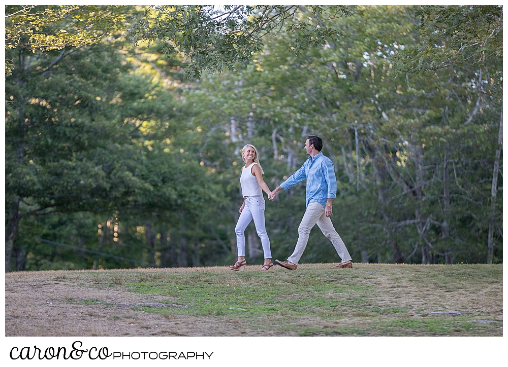 a woman wearing white, and a man in blue and tan, are walking together in a park, the woman is ahead of the man