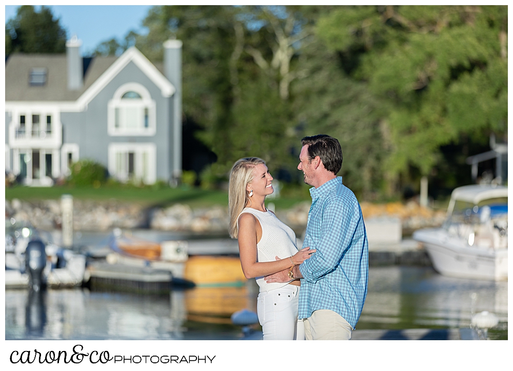 A woman in white, and a man wearing a blue shirt and tan pants, are standing face to face on a dock during their Boothbay Harbor engagement session in Boothbay Harbor, Maine