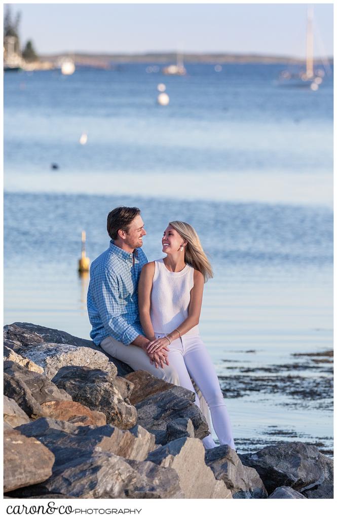 A woman and man are sitting on the rocks during a Boothbay Harbor, Maine engagment session