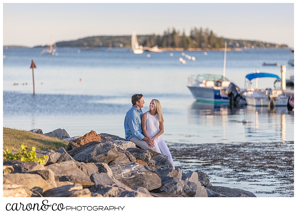 A woman and man sit together on the rocks with boats in the background of their Boothbay Harbor engagement photos