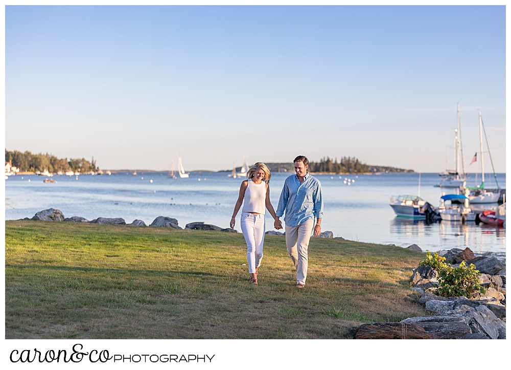 A woman in white, and a man in blue and tan, are walking hand in hand near Boothbay Harbor, during their Boothbay Harbor engagement photo session